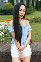 hermosa joven asiático niña en un verano azul blusa y blanco pantalones cortos es posando cerca el callejón de flores foto