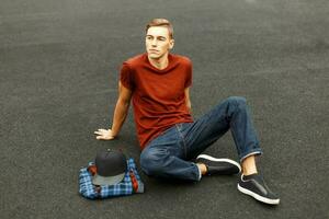 Handsome young man in shirt, jeans and sneakers sits on the pavement. photo