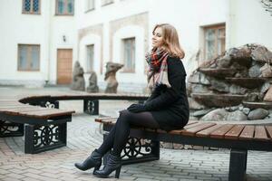 Young stylish girl in a scarf and black coat sitting on a bench on background of the building photo