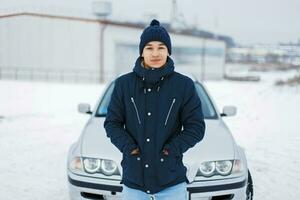 Handsome young man in winter jacket and knitted hat stands near the car photo