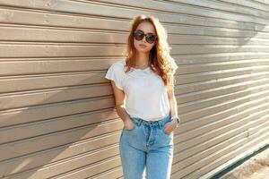 Vintage portrait of a beautiful young girl in a white T-shirt with sunglasses standing near a wooden wall on a sunny day. photo