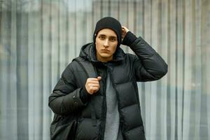 Handsome young man in a black hat and a warm jacket with a backpack posing outside of glass storefronts photo