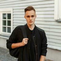 Handsome young man with hairstyle in a jacket with a backpack near a wooden house. photo