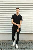 joven hermoso hombre en negro elegante vestir y blanco zapatillas posando cerca un ligero de madera pared. foto