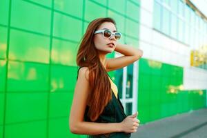 Stylish beautiful young woman in sunglasses and fashionable clothes stands near a green building. photo