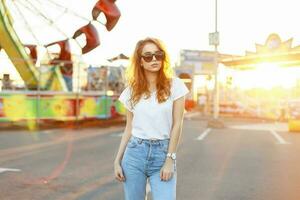 bonito joven mujer con Gafas de sol en un blanco camisa y Clásico pantalones caminando en el diversión parque a hermosa puesta de sol foto