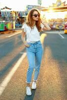 hermosa joven hipster niña en Gafas de sol en un blanco camiseta, Clásico pantalones y blanco zapatillas caminando en el diversión parque a puesta de sol. foto