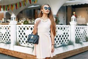 Fashionable beautiful woman in sunglasses, pink dress with a bag near a white fence photo