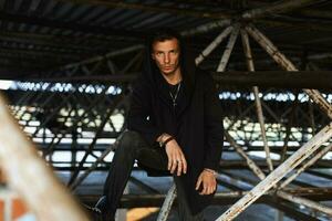 Stylish young man in a hood in black robes is posing on a background of metal pipes photo