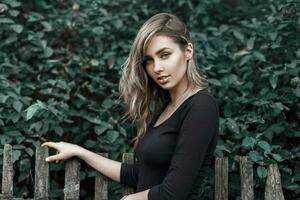 Beautiful girl in a black shirt standing near the wooden fence at the background of green foliage. photo