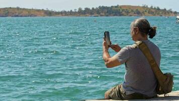 viajero hombre capturar Oceano ver con célula teléfono sentado en de madera muelle video