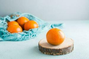 Ripe tangerine on a cut of a tree and tangerines in a mesh bag on a blue background. Eco-friendly fruits photo