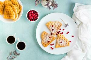 Heart shaped Belgian waffles with red berries and powdered sugar on a plate and two cups of coffee on the table. Valentine's Day. Top view photo