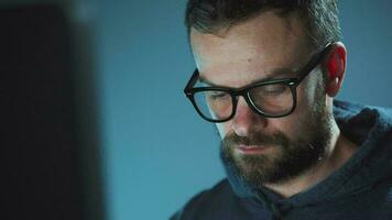 Bearded male hacker in hoodie and glasses working on a computer in a dark office room. Cybercrime concept video