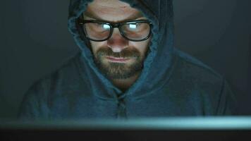 Male hacker in the hood and glasses working on a computer in a dark office room. Cybercrime concept video