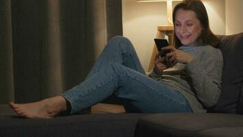 leende kvinna liggande på de soffa i en mysigt rum och använder sig av smartphone för surfing internet i de kväll. avslappning och livsstil teknologi video