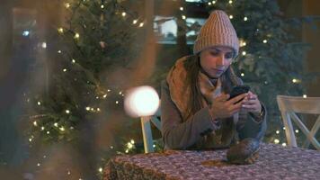 donna si siede nel un all'aperto bar terrazza nel inverno e chat nel sociale reti, utilizzando sua smartphone. Natale decorazioni su il sfondo. comunicazione, Natale e nuovo anno vacanze concetto video