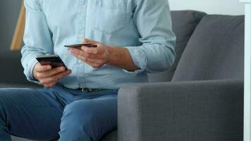 Formally dressed man sits on a sofa and enters a credit card number into a smartphone to pay online. Online shopping video