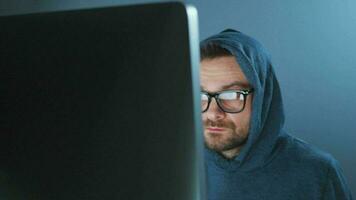 Male hacker in the hood and glasses working on a computer in a dark office room. Cybercrime concept video