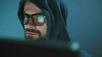 Male hacker in the hood and glasses working on a computer in a dark office room. Cybercrime concept video
