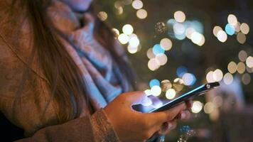 Close-up of woman uses a smartphone in winter on the background of a Christmas tree decorated with lights. Communication, Christmas and New Year Holidays concept video