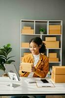 Startup small business SME, Entrepreneur owner African woman using smartphone or tablet taking receive and checking online purchase shopping order to preparing pack product box. in office photo