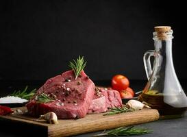 Raw piece of beef with spices pepper, rosemary sprig, salt and olive oil on a wooden board, black background photo