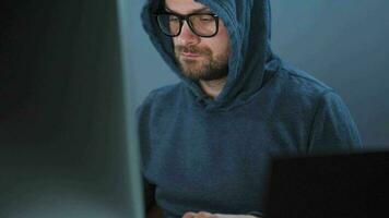 Male hacker in the hood and glasses working on a computer in a dark office room. Cybercrime concept video