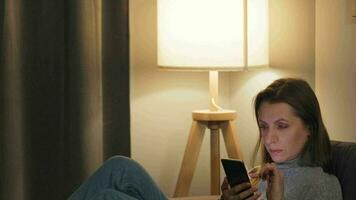 mujer acostado en el sofá en un acogedor habitación y utilizando teléfono inteligente para surf Internet en el noche. en línea compras, estilo de vida tecnología video