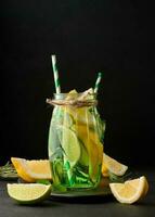Lemonade in a transparent glass with lemon, lime, rosemary sprigs and mint leaves on a black background photo