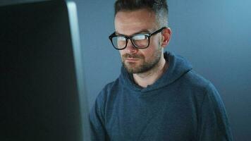 Bearded male hacker in hoodie and glasses working on a computer in a dark office room. The monitor screen is reflected in the glasses. Cybercrime concept video