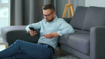 barbado hombre en lentes se sienta en el alfombra cerca el sofá y hace pago vía el Internet con un crédito tarjeta y teléfono inteligente en línea compras, tienda en el sofá, estilo de vida tecnología video