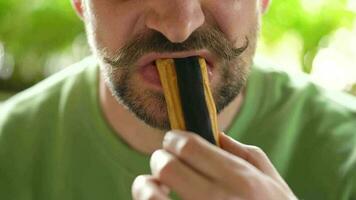 Bearded man with curled up mustache eats chocolate eclair in cafe. Close-up video