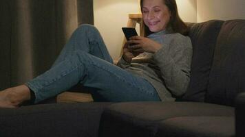 leende kvinna liggande på de soffa i en mysigt rum och använder sig av smartphone för surfing internet i de kväll. avslappning och livsstil teknologi video