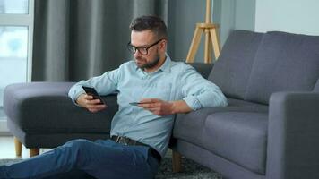 Bearded man in glasses sits on the carpet near the sofa and makes payment via the Internet with a credit card and smartphone. Online shopping, shop on the couch, lifestyle technology video