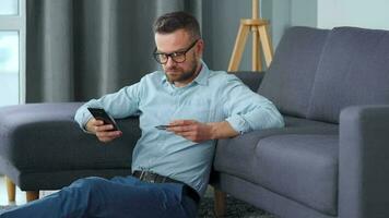 barbado hombre en lentes se sienta en el alfombra cerca el sofá y hace pago vía el Internet con un crédito tarjeta y teléfono inteligente en línea compras, tienda en el sofá, estilo de vida tecnología video