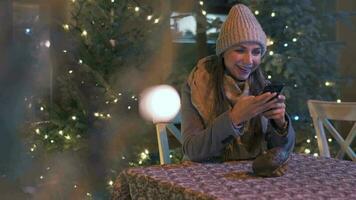 vrouw zit in een buitenshuis cafe terras in winter en chatten in sociaal netwerken, gebruik makend van haar smartphone. Kerstmis decoraties Aan de achtergrond. communicatie, Kerstmis en nieuw jaar vakantie concept video