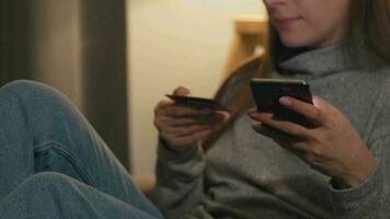 vrouw aan het liegen Aan de sofa in een knus kamer en maakt een online aankoop gebruik makend van een credit kaart en smartphone. online winkelen, levensstijl technologie video