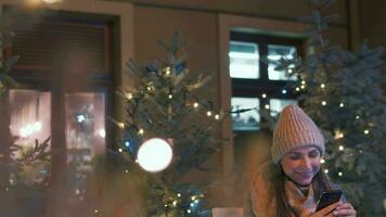 mujer se sienta en un al aire libre café terraza en invierno y chateando en social redes, utilizando su teléfono inteligente Navidad decoraciones en el antecedentes. comunicación, Navidad y nuevo año Días festivos concepto video