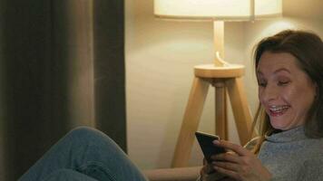 Smiling woman lying on the sofa in a cozy room and using smartphone for surfing internet in the evening. Relaxation and lifestyle technology video