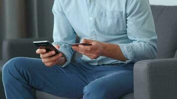 Formally dressed man sits on a sofa and enters a credit card number into a smartphone to pay online. Online shopping video