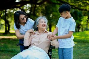 Happy senior Asian grandmother uses wheelchair with her daughter and grandchild in park, Grandson came to visit elderly grandmother and hold hand. Concept of happy family, good relationship together photo