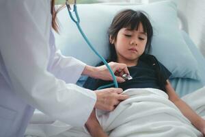 Asian woman pediatrician doctor hold stethoscope for exam a little girl patient and heck heart lungs of kid, Good family doctor visiting child at home, Healthcare and medicine for childhood concept. photo