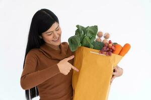 Happy Asian woman smiling and point the finger to a paper bag and carries a shopping bag after the courier from grocery came to deliver at home. Concept of Supermarket delivery for a new lifestyle. photo