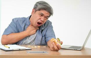 Asian man working and eating a burger on office desk and holding his neck after choking foods. Concept of a busy businessman cannot work-left balance and not taking care of health Eat only junk food photo