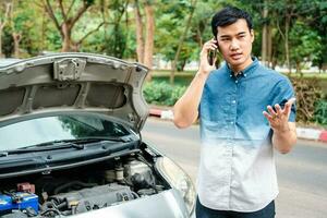 hombre asiático enojado y usando un teléfono móvil pidiendo ayuda después de una avería en el coche en la calle. concepto de problema del motor del vehículo o accidente y ayuda de emergencia de un mecánico profesional foto