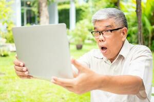 Portrait of an old elderly Asian man is holding a computer laptop and surprised after got some email and read some news in the backyard. Concept of no Ageism and Hobbies after retirement. photo