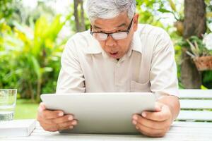 Portrait of an old elderly Asian man is holding a computer laptop and surprised after got some email and read some news in the backyard. Concept of no Ageism and Hobbies after retirement. photo