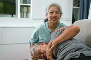 Asian old elderly man is In pain with his hands on his chest and having a heart attack in the Living room and his wife is shocked and confused, helpless. Concept of healthcare and Elderly caregivers photo