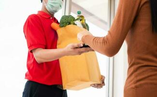 Asian delivery man holding holding a bag of fresh food for giving to customers and holding smartphone for Receive payments at home. Concept of express grocery service and new lifestyle photo